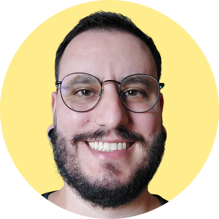 Foto de Fernando Fantinel sorrindo usando óculos de grau, alargadores pretos, camiseta preta e barba em fundo amarelo.